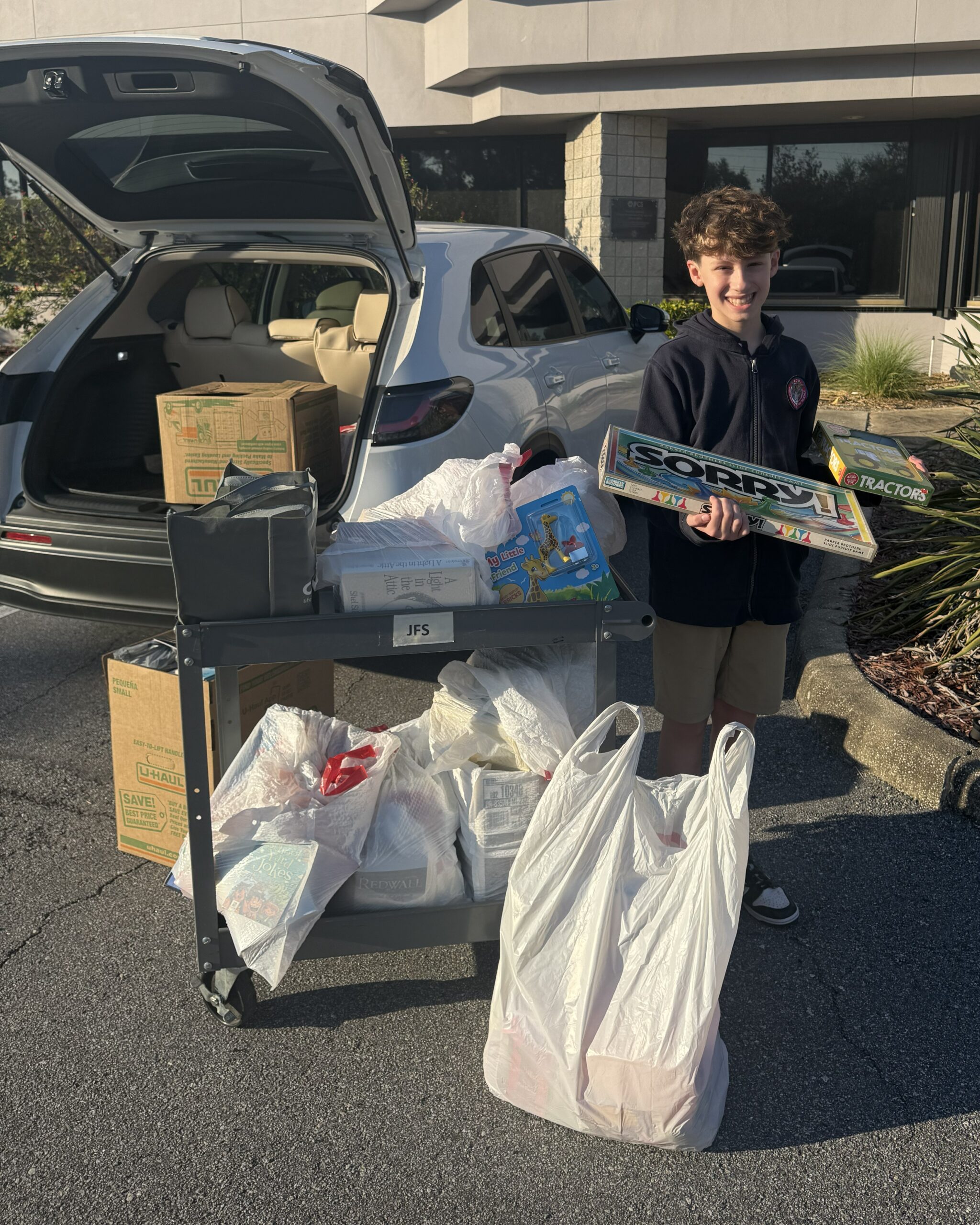 Young boy stands holding a game of 'Sorry' next to an open car with it's trunk open. There are several bags and boxes of games and books surrounding him. 