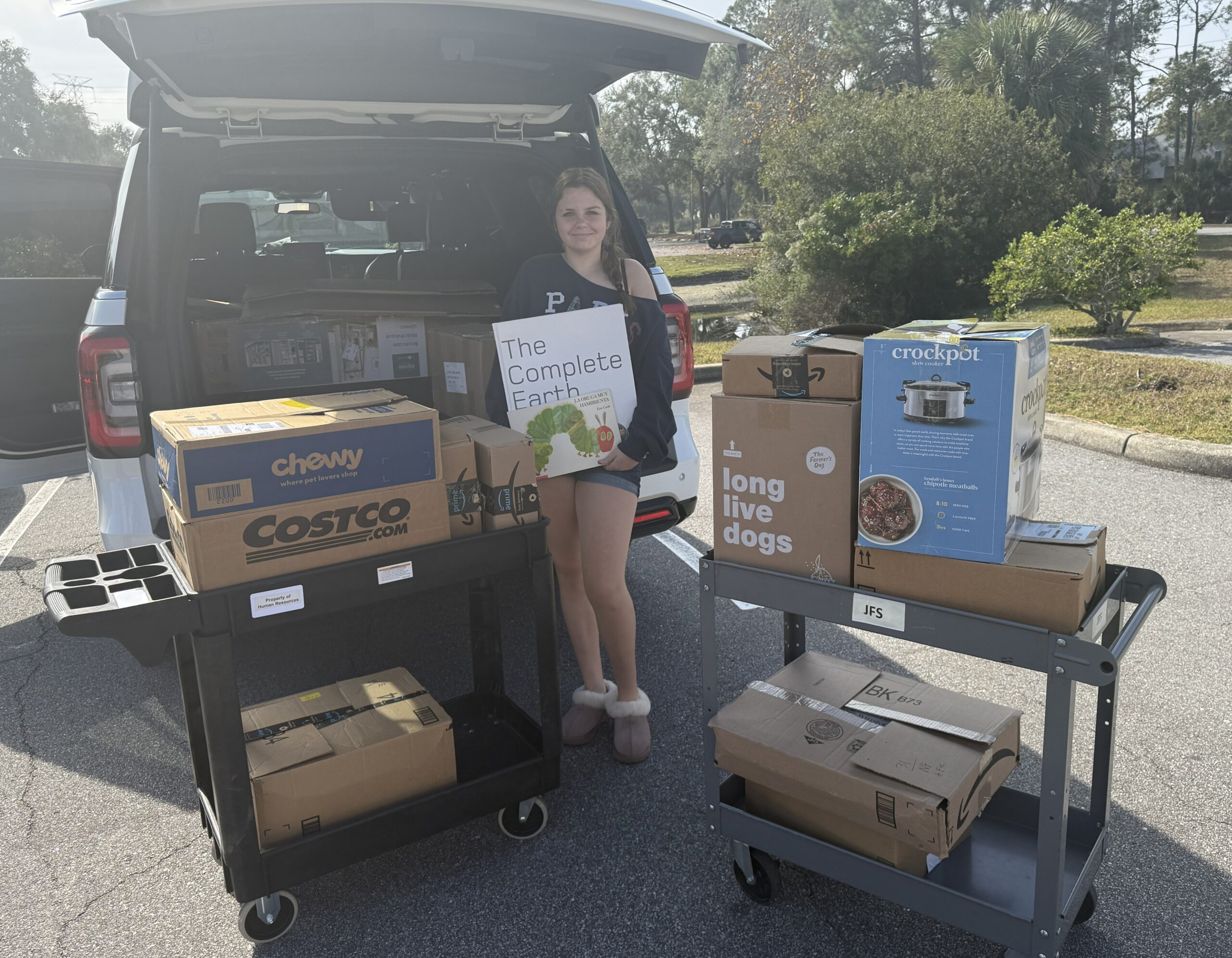 Girl stands next to car with an open trunk. She holds two books, smiling, while surrounded by boxes of other books.